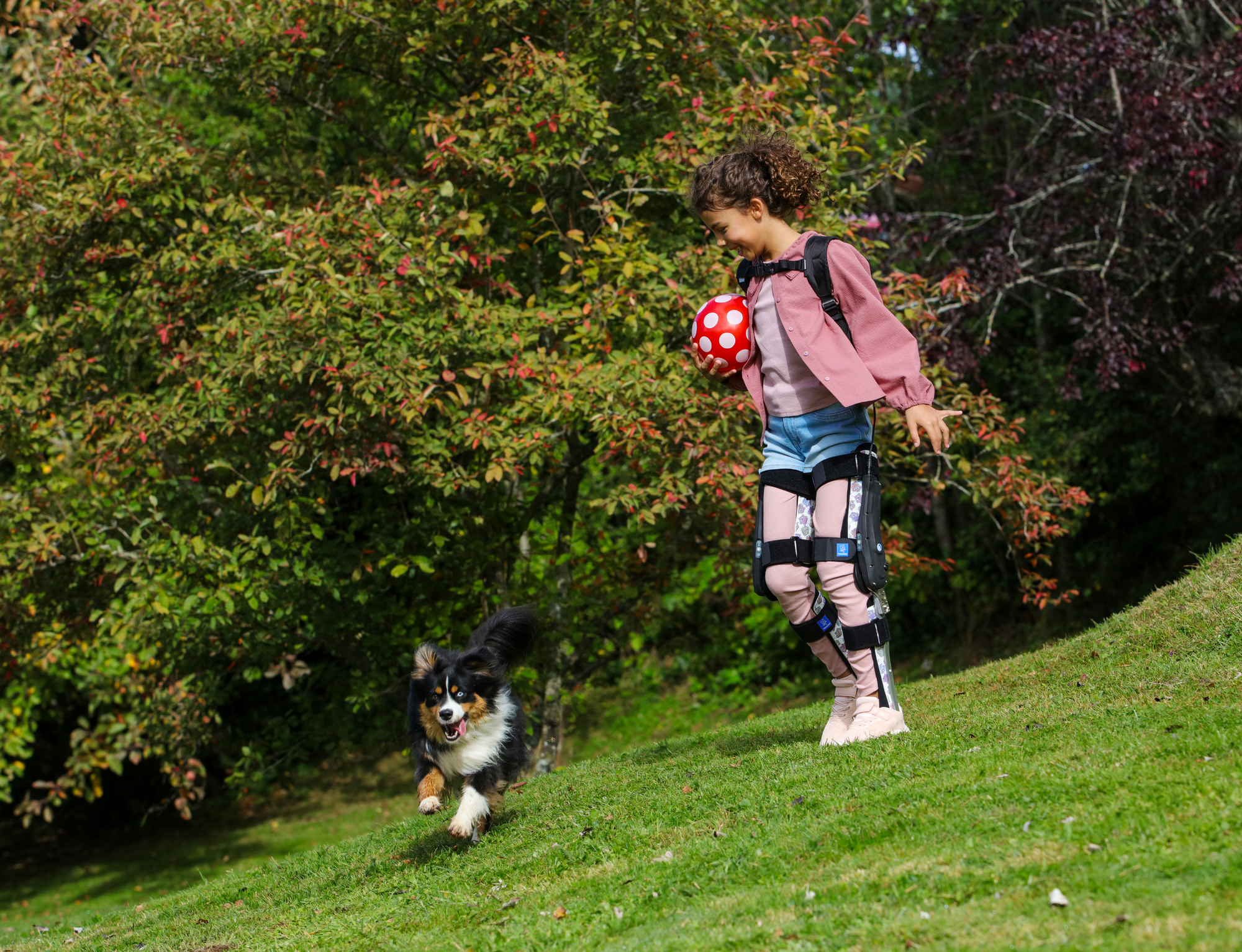 A young child wearing leg braces walks down a grassy hill while holding a red polka-dot ball, smiling at a small dog running beside them. Trees and greenery fill the background, creating a lively outdoor scen