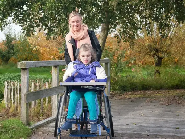 A picture of a woman pushing a girl in a wheelchair in a woodland park. 