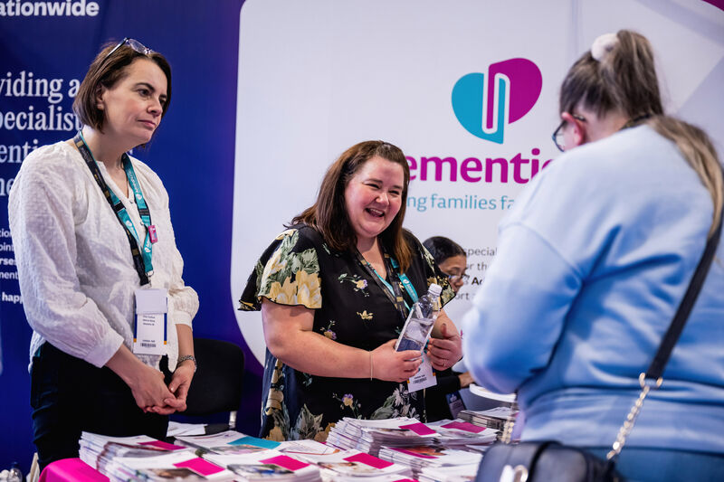 Exhibitors at a dementia-support stand smiling and speaking with a visitor, with brochures and branded materials displayed on the table.