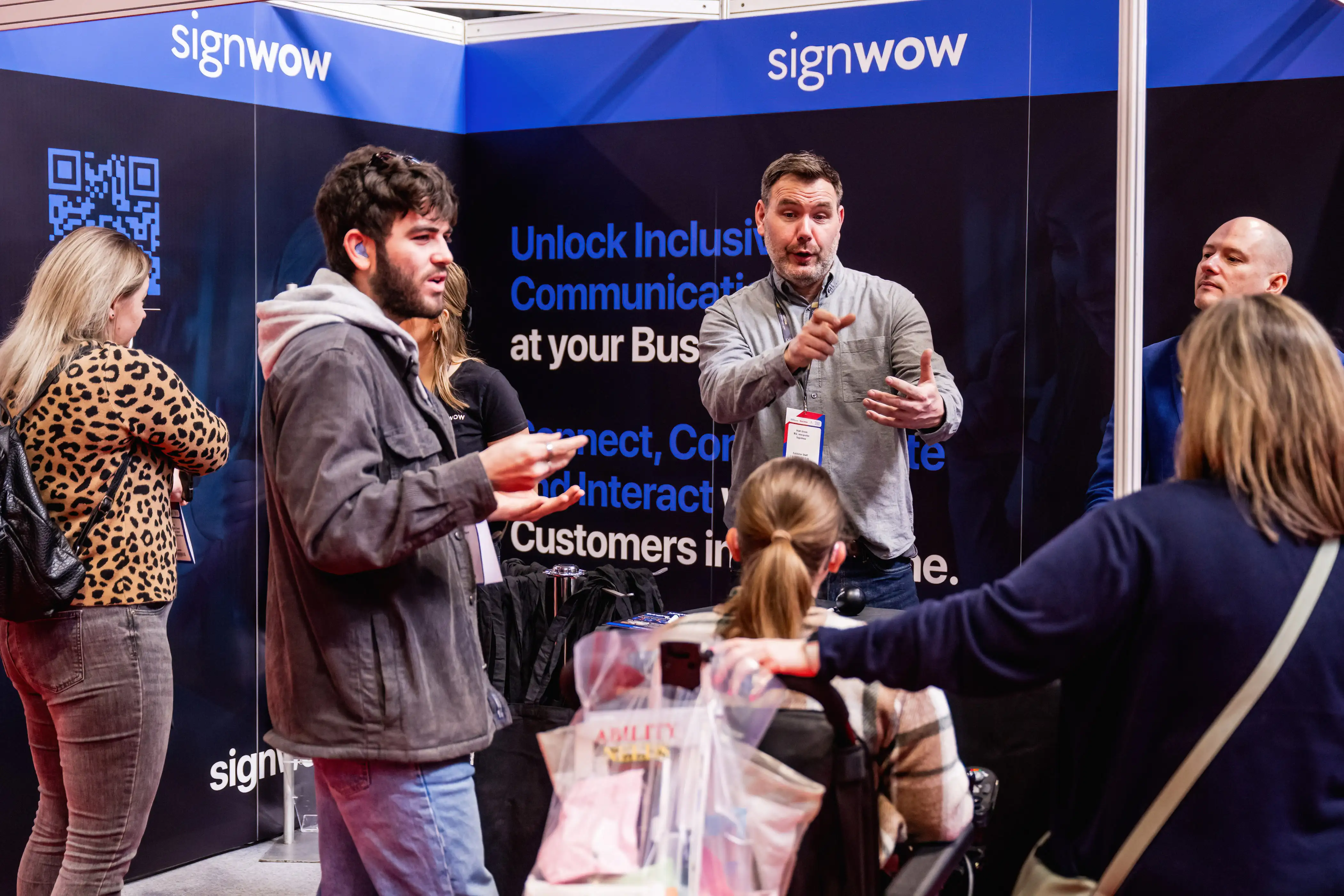 People engaging in conversation at the SignWow exhibition stand. A presenter gestures while explaining inclusive communication technology to visitors.