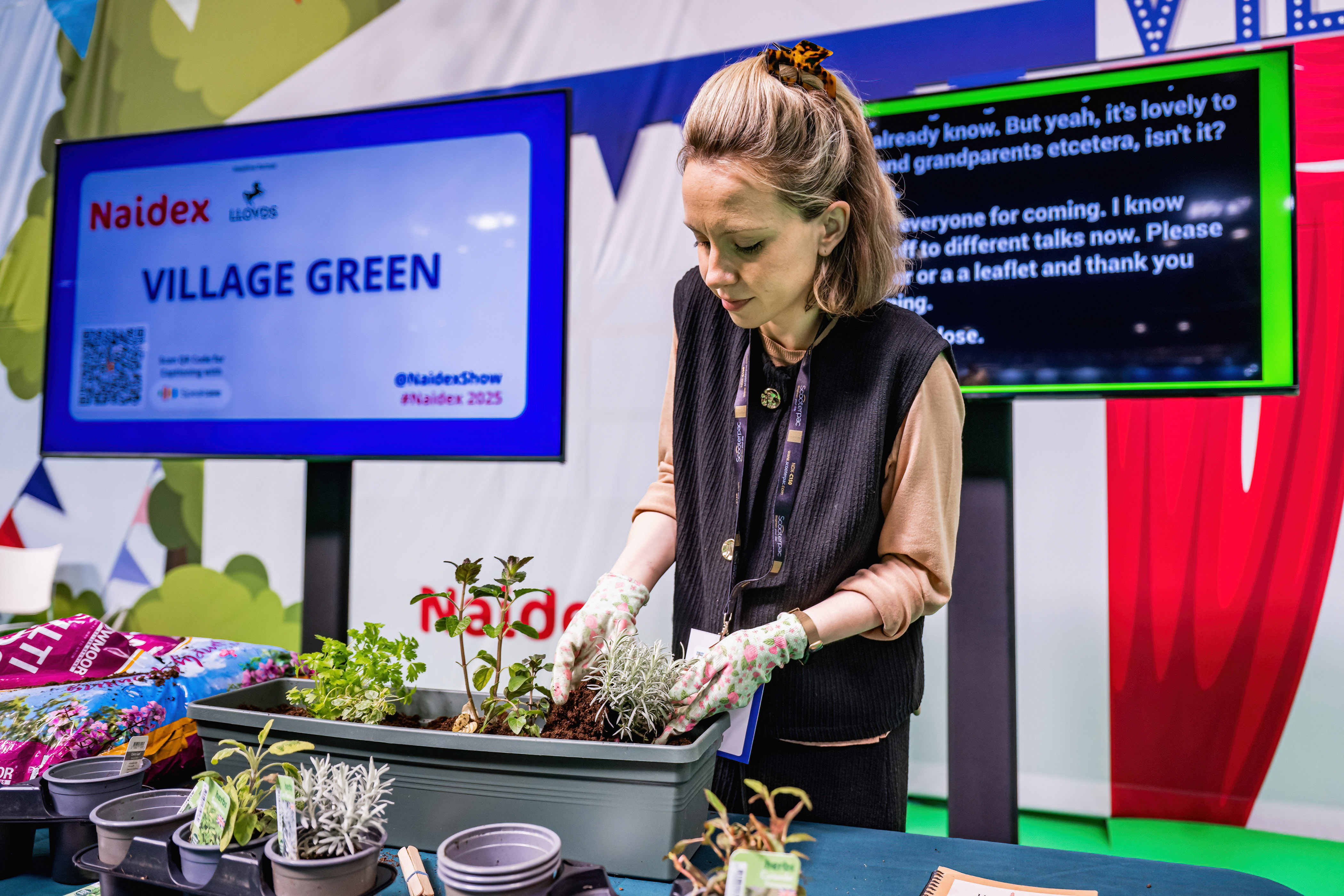 A photograph of an attendee participating in a hands-on gardening session at the Naidex "Village Green" feature area. The woman is wearing floral gardening gloves while potting plants in a grey window box. In the background, a large digital screen displays the Naidex and Lloyds partner logos alongside live-captioned text for accessibility.