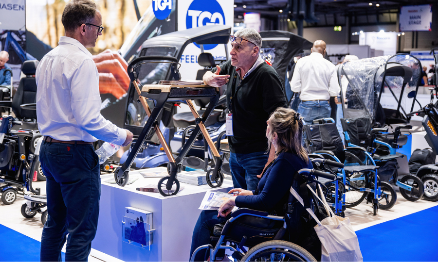 Three people are having a conversation, one person is an exhibitor and another is a wheelchair user. They are surrounded by various wheelchairs on display