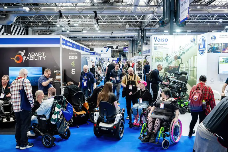 Visitors exploring accessibility products and mobility solutions at a busy exhibition hall, with several attendees using wheelchairs and mobility scooters.