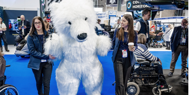 A woman is holding the arm of someone dressed in a big polar bear costume, they are at the Naidex event