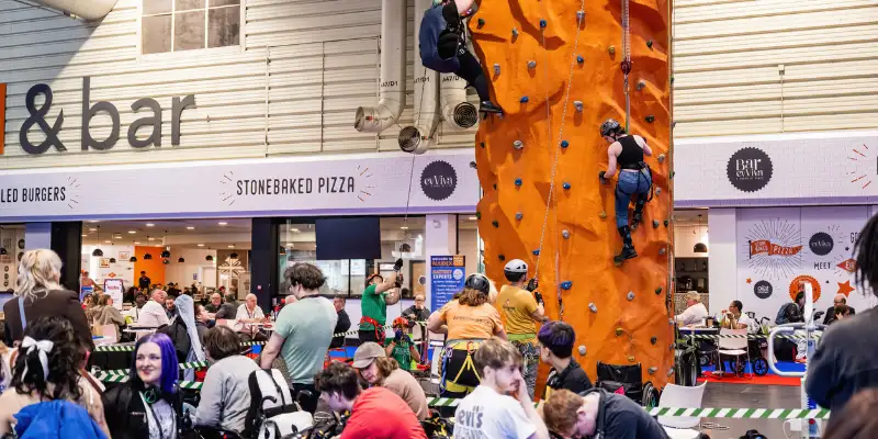 A wide shot picture of the orange adaptative climbing wall there are two people on the climbing wall, you can also see lots of different people around the show.