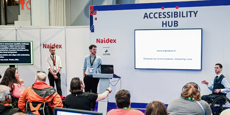 A man is presenting a talk at the accessibility hub, there is a big white screen and audience members in view.