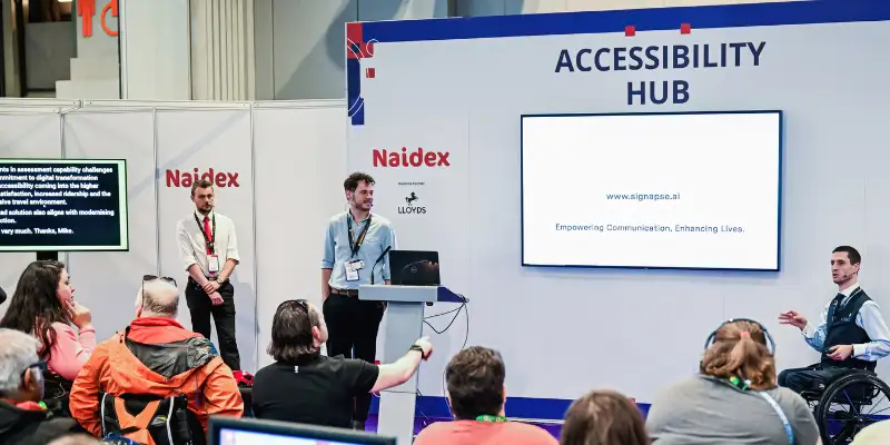 A man is presenting a talk at the accessibility hub, there is a big white screen and audience members in view.