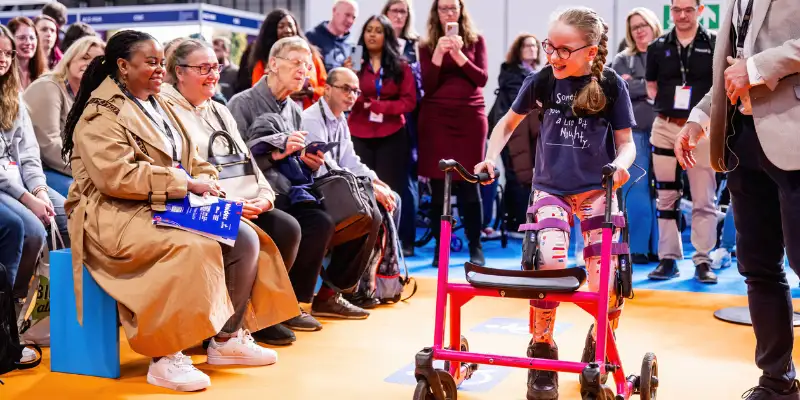 A girl is using an assistive walking frame, she is smiling and walking past the front row of an audience who is also smiling