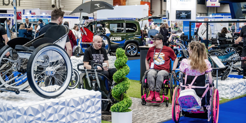 A picture of three wheelchair users talking at the event, there are lots of wheelchairs and vehicles on display
