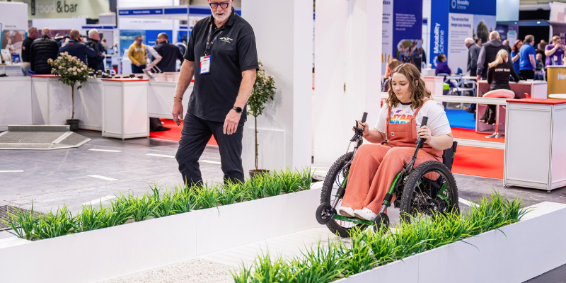 A girl is in a powerchair testing on the Mobility Test Track at Naidex, she is wearing light pink dungarees. The test track has small white walls along the track and at the top of them is a line of fake grass.
