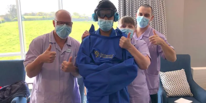 Four people are facing the camera, three are holding their thumbs up, the other has their arms crossed and is wearing goggles, a mask and headphones. The other three people are wearing masks as well.