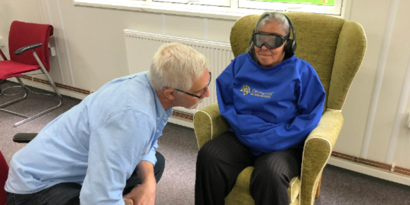 One person is sat on a chair, they are wearing goggles and headphones, they are also in a blue shirt which is designed to restrain their arms. Another person is knelt beside them.