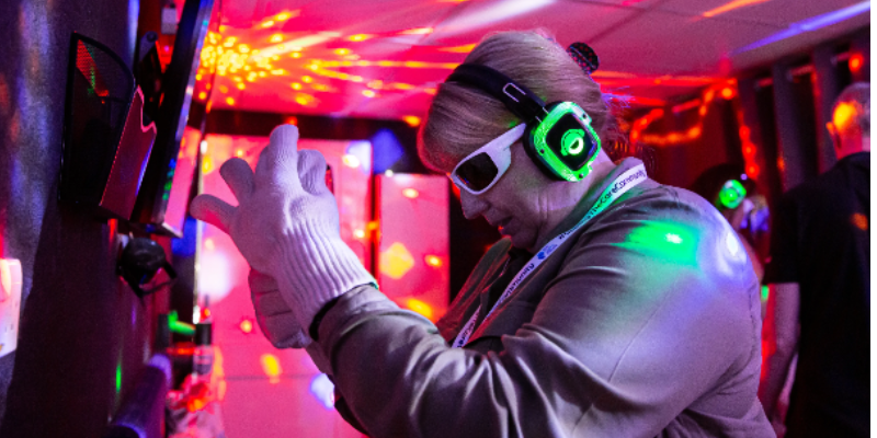 A person wearing headphones, sunglasses, and white gloves interacts with a wall in a room filled with colorful, flashing lights, creating an immersive, sensory-rich environment.