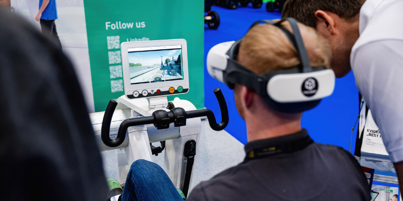 A person wearing a VR headset uses a stationary bike with a screen displaying a virtual outdoor scene, while another person observes nearby.