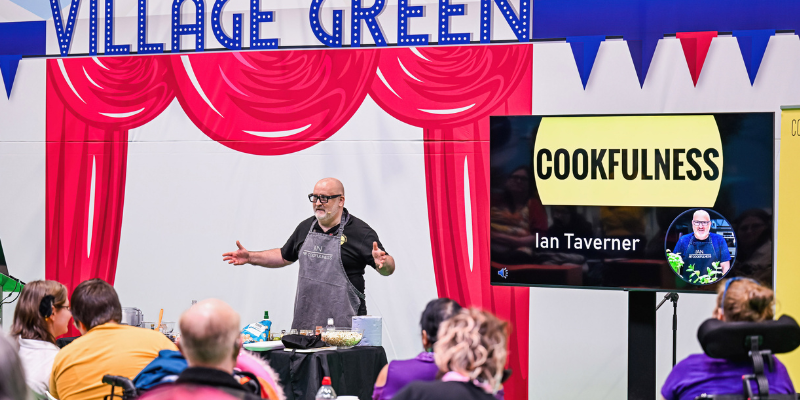 A man is addressing a crowd of people, he is on the village green and is wearing an apron, he is delivering cooking advice.