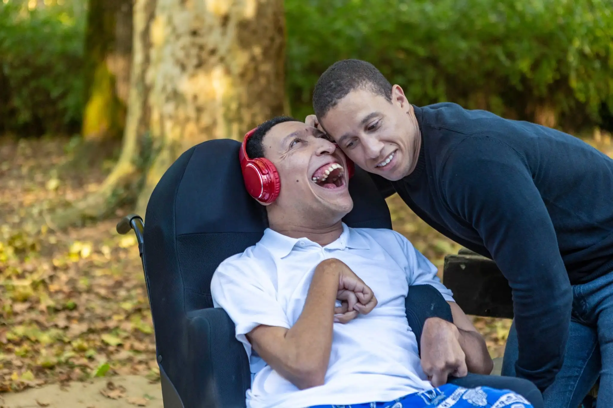 2 young people hugging each other. On the left is a wheelchair user who has cerebral palsy and wearing red headphones, on the right is a young man wearing a blue jumper.  