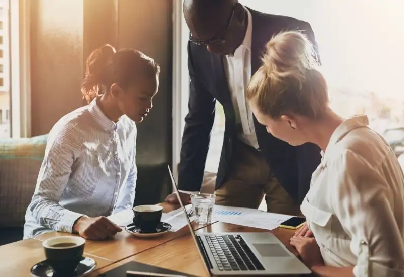 Photo of 3 people working in an office all looking at a spreadsheet. There are 2 women with a man in the middle.
