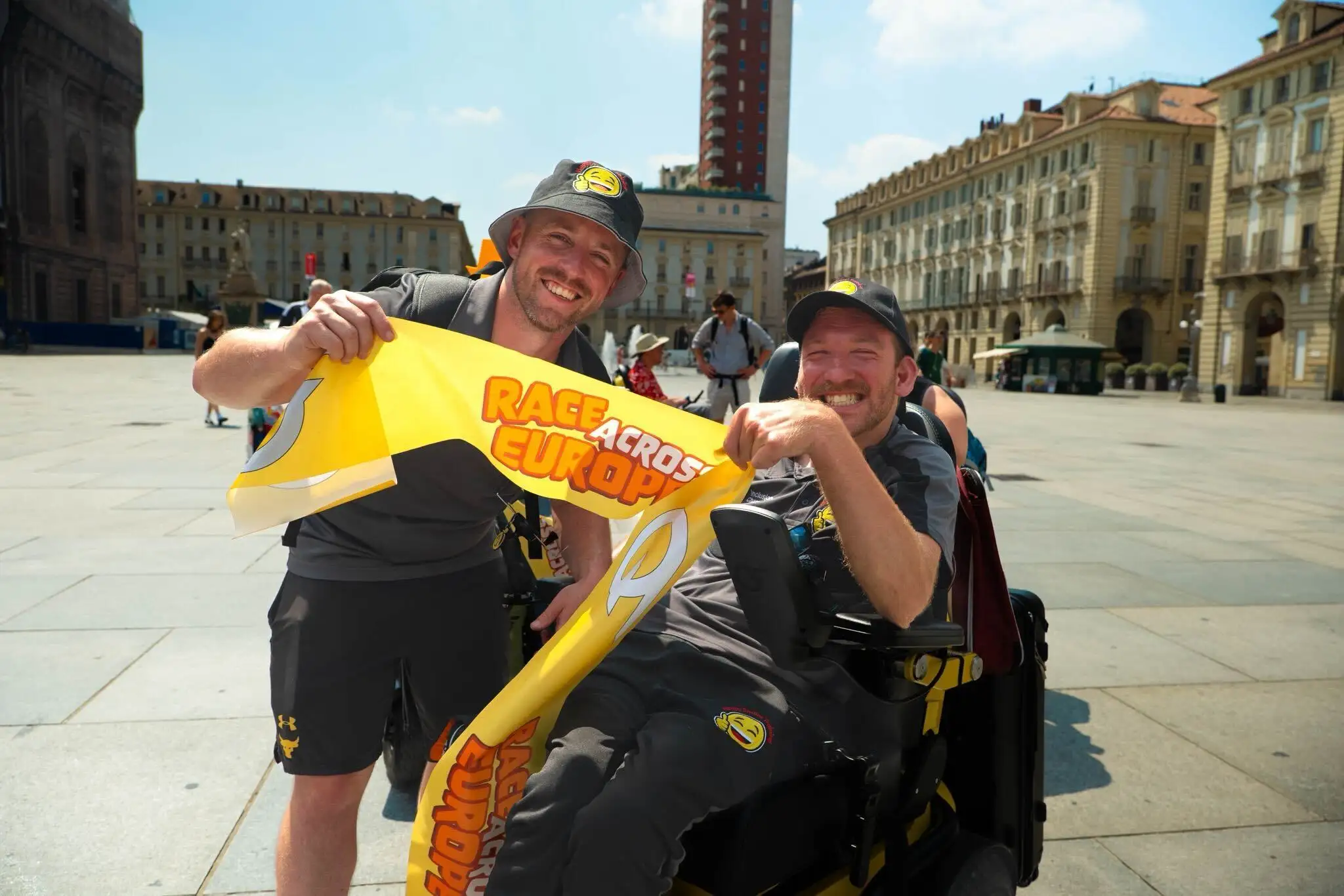 A picture of two people smiling into the camera, one is in a wheelchair and they are each holding a side of a yellow scarf that says "Race Across Europe" in orange writing 