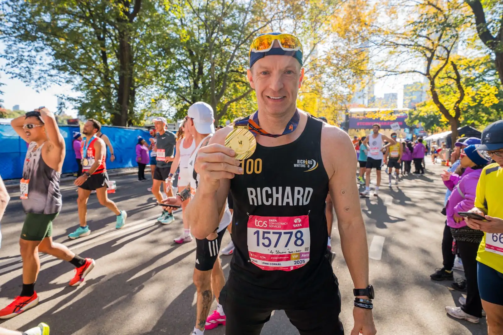 A picture of Richard Whitehead at the end of the New York marathon, he is smiling and holding his medal up to show the camera. It is sunny and there are lots of runners and people gathered around