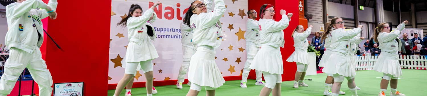 A dance group, wearing all white are performing a dance at Naidex on the village green.