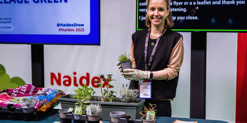 A woman is smiling at the camera as she stands behind a desk, the desk has different plants on it and she is holding one up to show the camera.