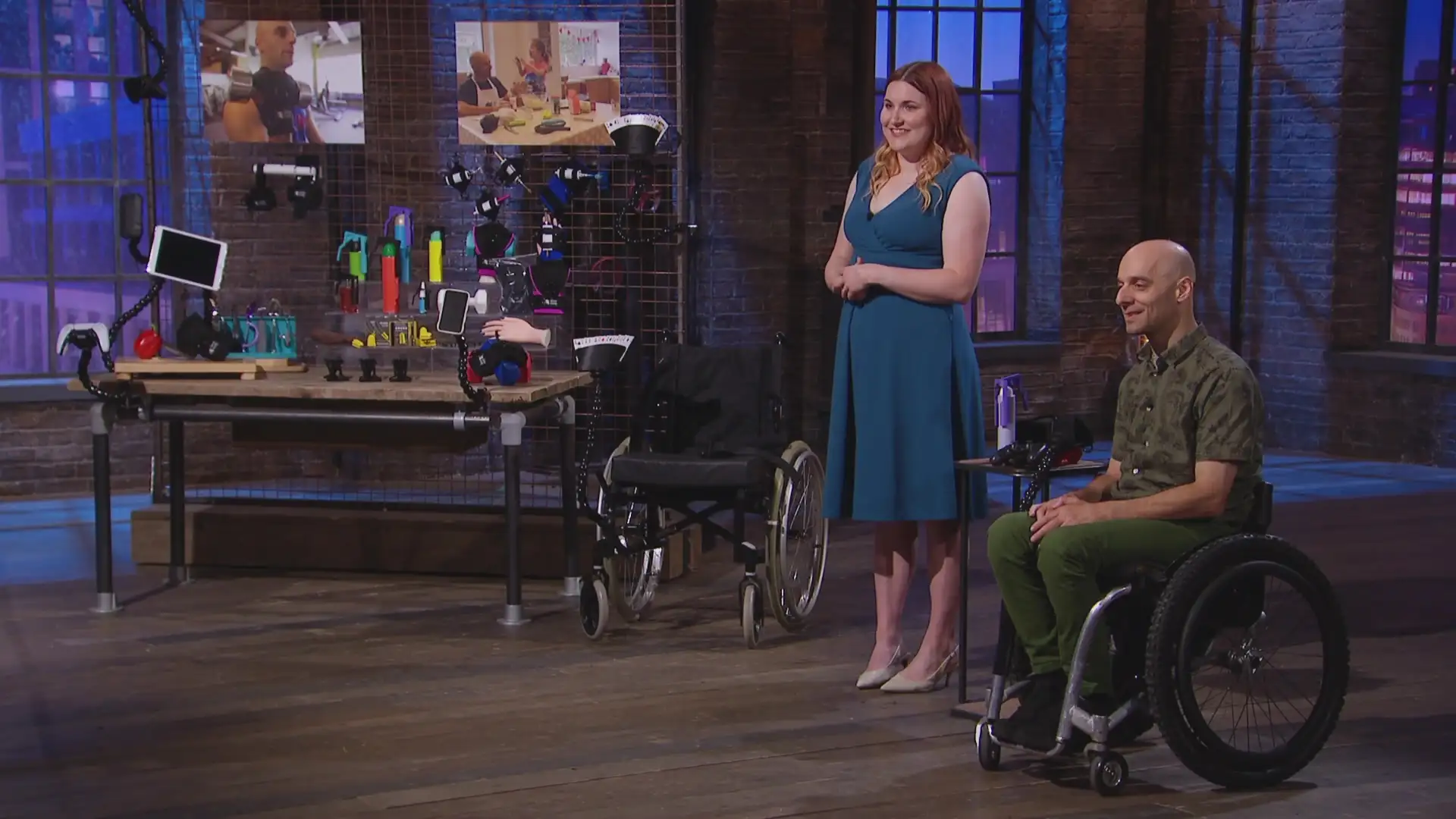 Two people present assistive technology in a studio setting, Dragons Den. A woman in a teal dress stands beside a man seated in a wheelchair, both smiling. Behind them is a display of adaptive devices and tools on a workbench, with screens showing demonstrations. The space has an industrial-style interior with brick walls and studio lighting.