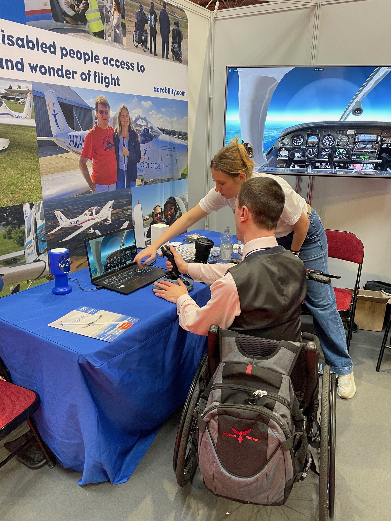 A wheelchair user tries a flight simulator at an exhibition stand, guided by a staff member pointing at a laptop screen, with aviation visuals displayed and Aerobility branding promoting accessible flying.
