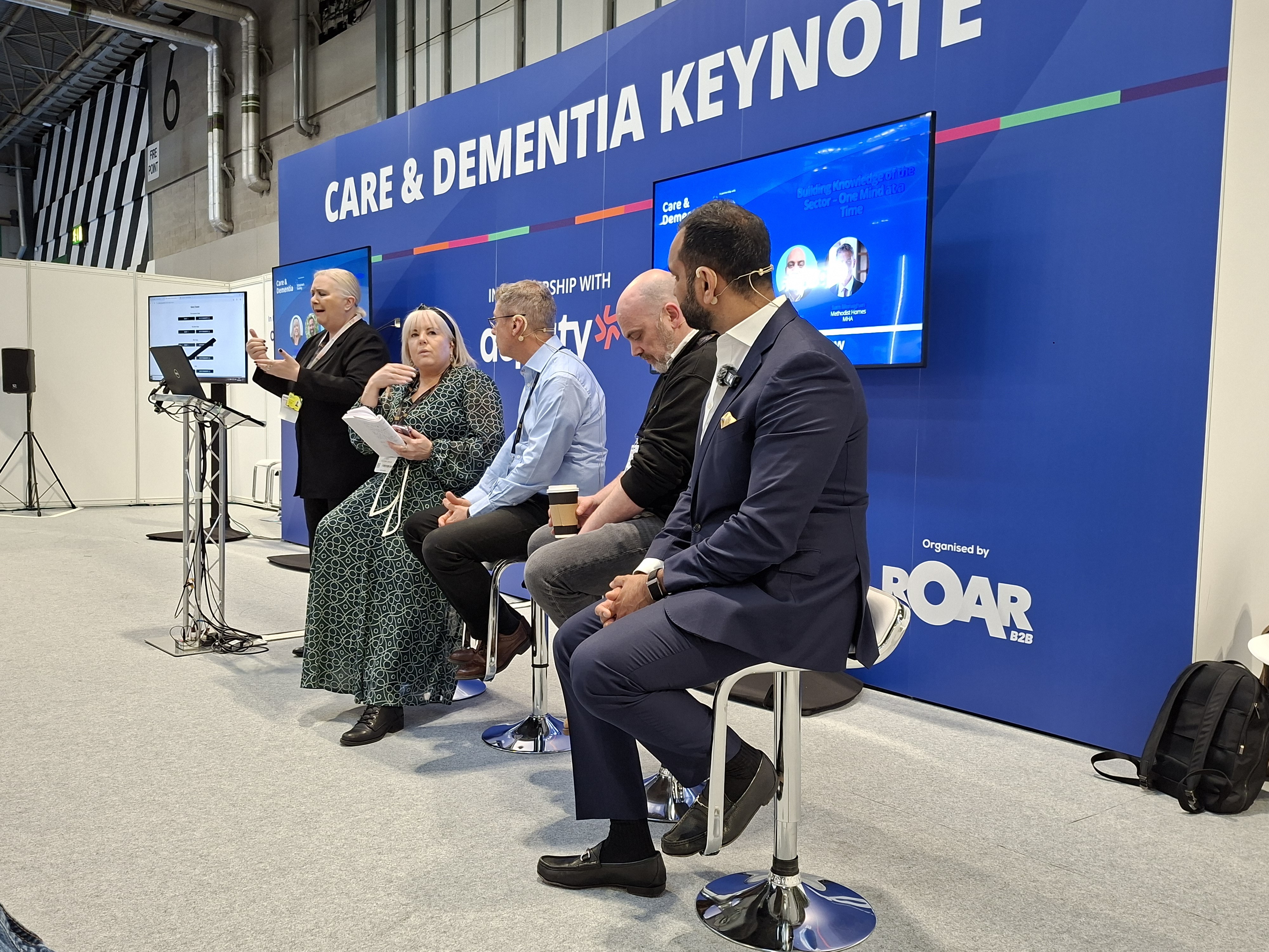 Panel discussion taking place on a Care & Dementia keynote stage, with multiple speakers seated on stools while one presenter speaks at a podium. The backdrop features event branding, partner logos, and screens displaying session content.