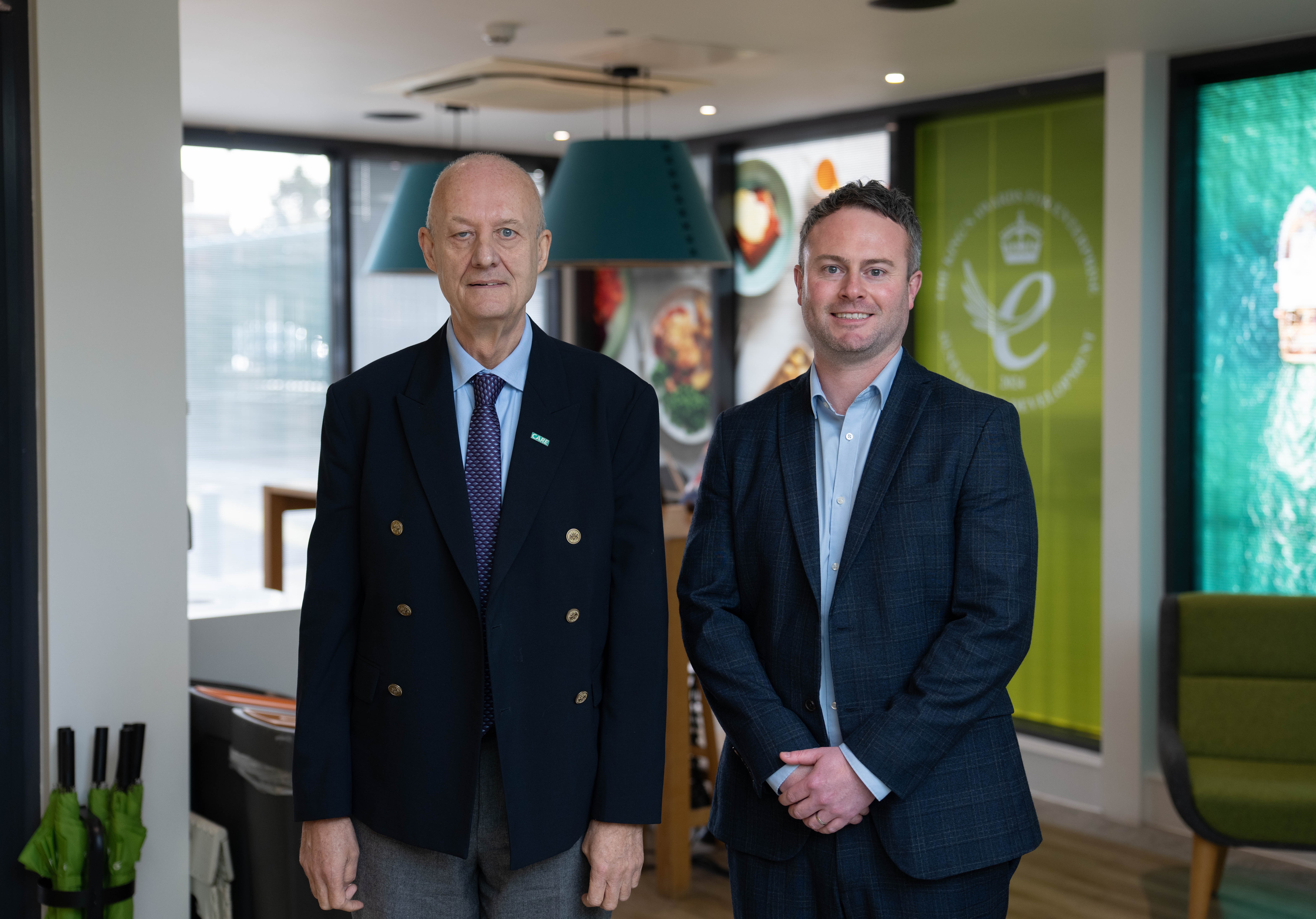 Two professionally dressed men standing side by side in a modern indoor setting, facing the camera. The space appears to be a contemporary lounge or office with large windows, pendant lighting, and branded décor in the background.