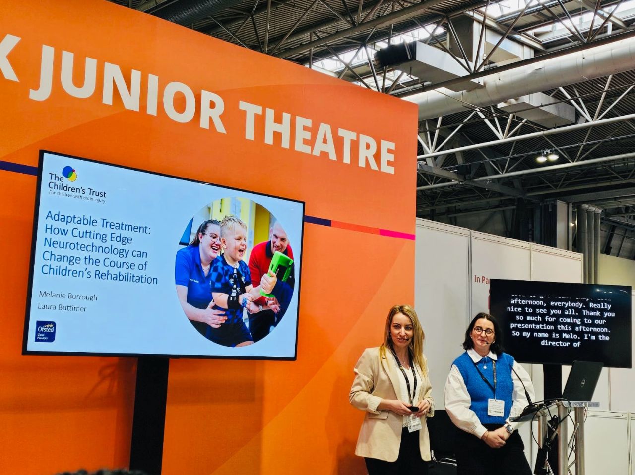Two presenters stand at a podium in a “Neuro Junior Theatre” area, delivering a talk on neurotechnology and children’s rehabilitation, with a large screen displaying their presentation and event branding in the background.