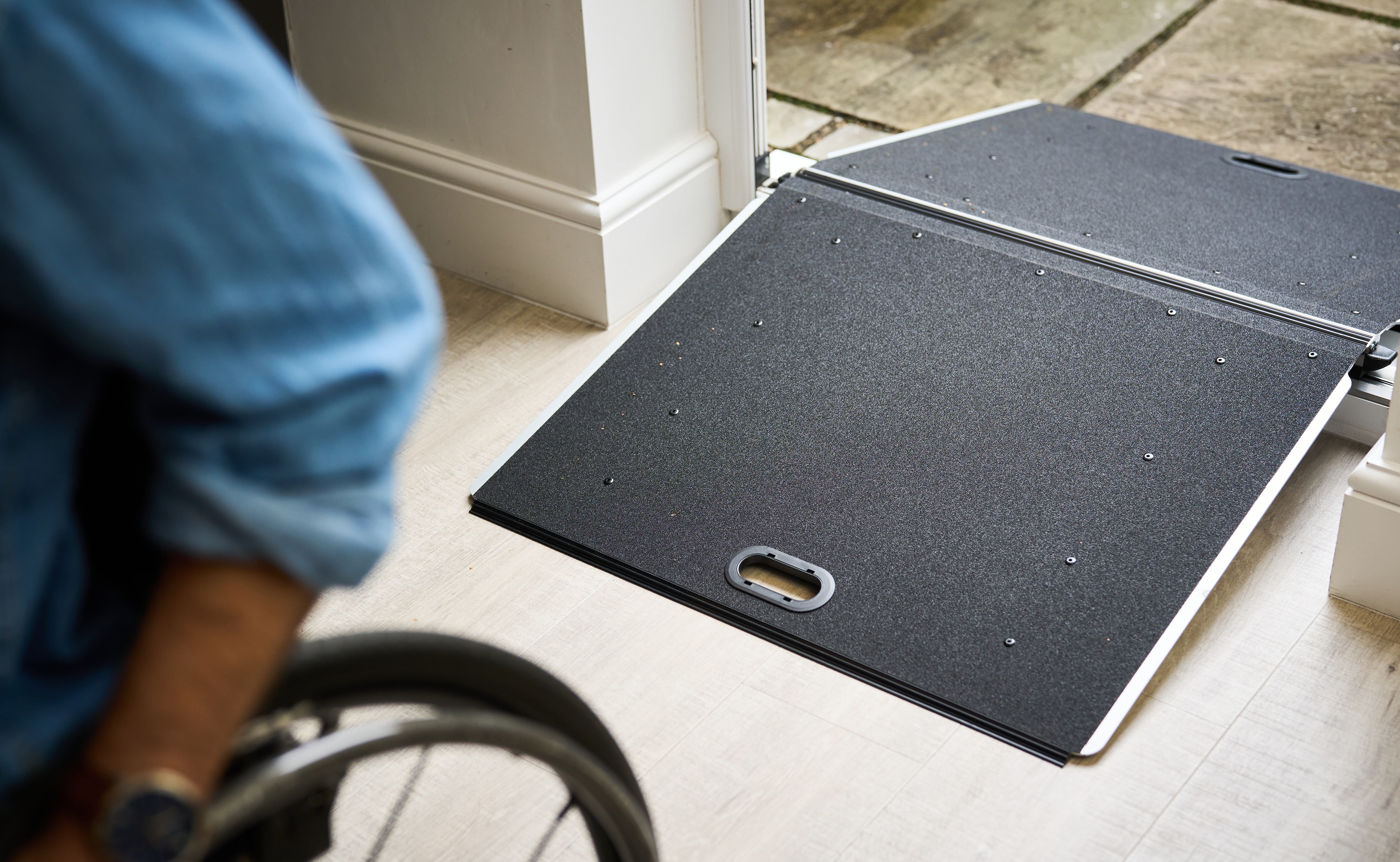 A portable ramp is placed over a doorway threshold to provide wheelchair access. In the foreground, part of a wheelchair and a person’s arm are visible, approaching the ramp from inside a home.