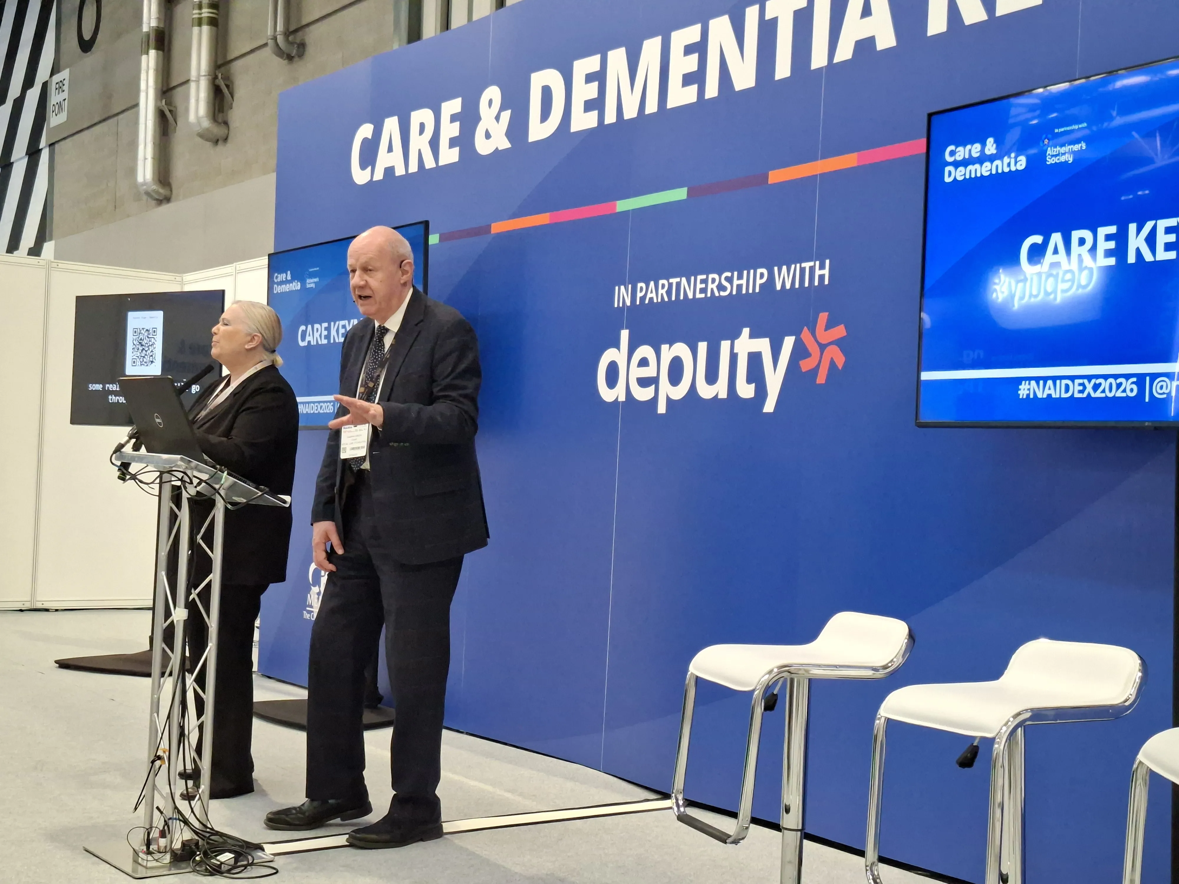 Damien Green and another speaker presenting on stage at a Care & Dementia event, standing beside a podium with a laptop. Behind them is a large branded backdrop reading “Care & Dementia” and “in partnership with deputy,” along with screens displaying event content and hashtags.