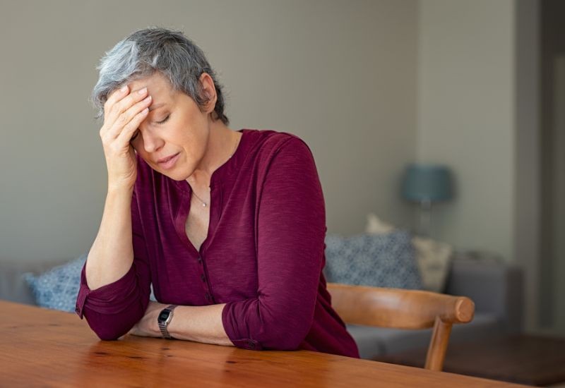 A middle-aged woman with short grey hair sits at a wooden table, resting her head in her hand and looking down, appearing stressed or fatigued. The background shows a softly lit home setting with a sofa and lamp.