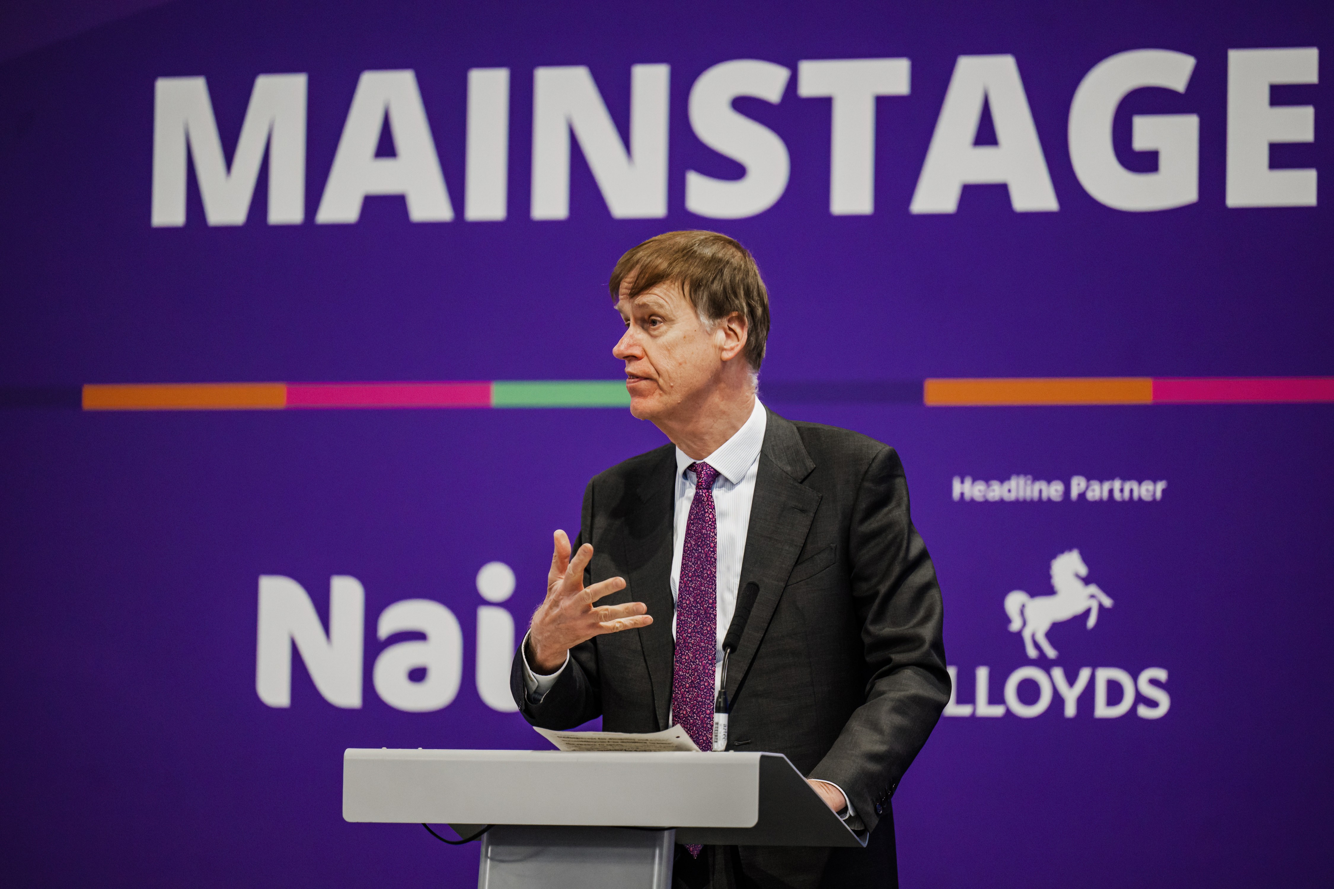 A speaker in a suit delivers a presentation at a podium on a main stage, with a large purple backdrop displaying “MAINSTAGE” and Naidex branding behind him.