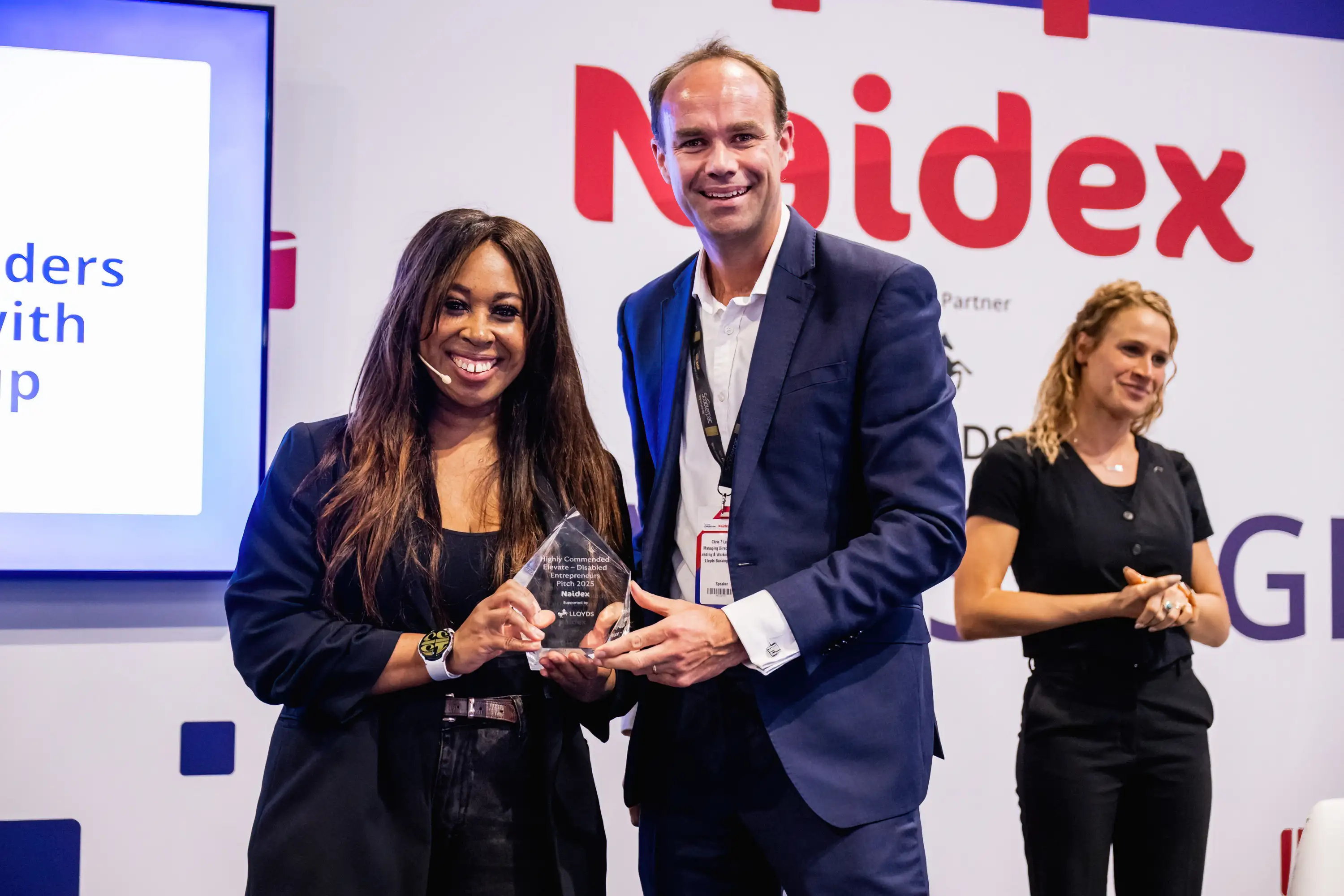 A picture of two people from the Elevate 2025 awards. They are smiling looking into the camera, they are both holding a glass award,. 