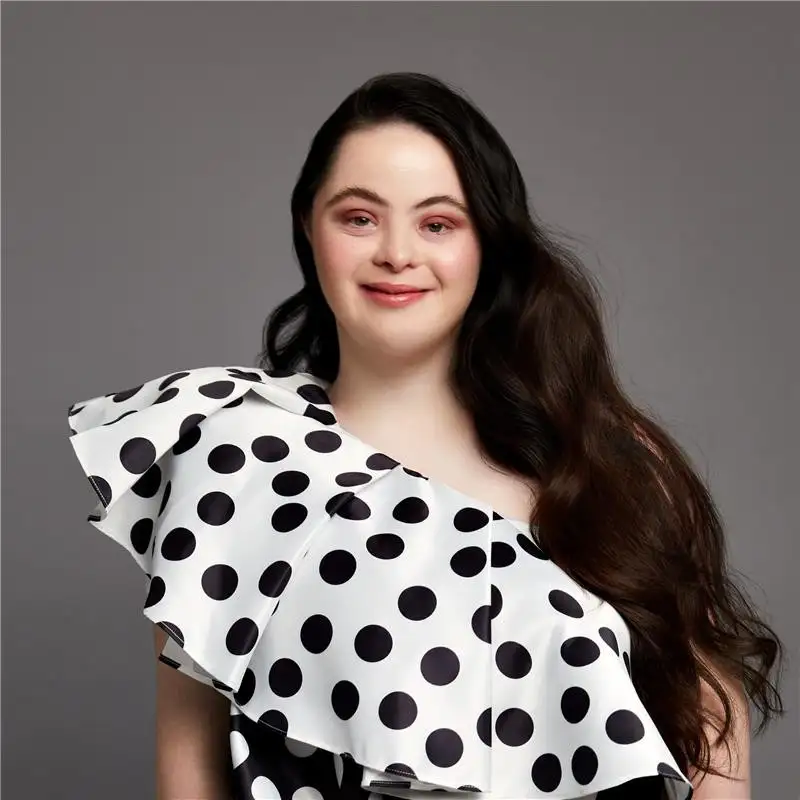 A studio portrait of Ellie Goldstein with long dark hair, wearing a white one-shoulder top with black polka dots. She is facing the camera against a plain grey background.