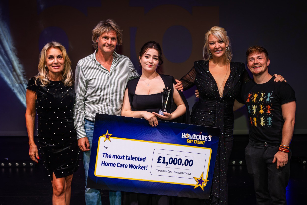 Macey Shaw, winner of the Homecare's Got Talent award, stands centre stage holding a glass trophy and a large novelty cheque for £1,000 made out to 'The most talented Home Care Worker.' She is flanked by four presenters, two women in sparkly black dresses and two men, all smiling with their arms around her in celebration.