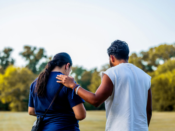 Two people walking side by side in a park, with one placing a supportive hand on the other’s shoulder, surrounded by greenery.