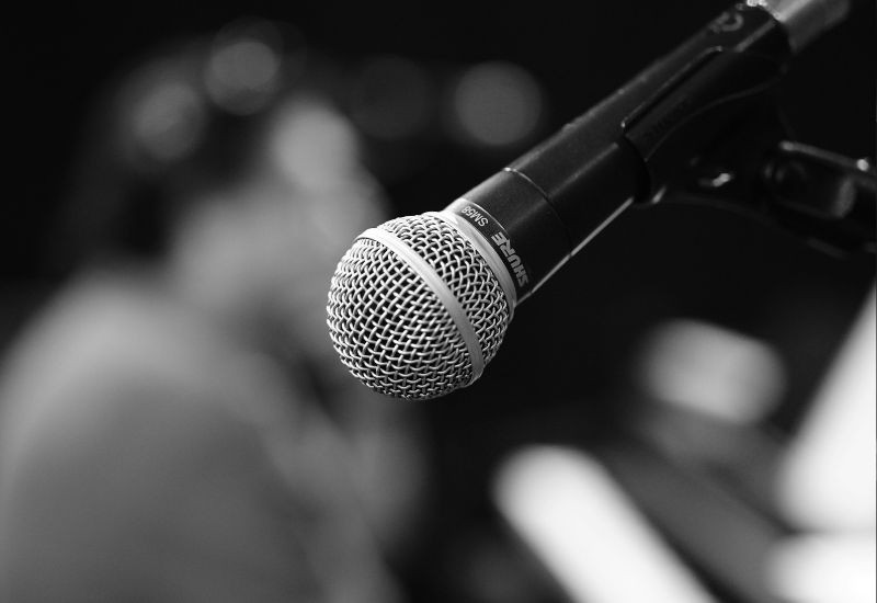 Alt: A close-up of a handheld microphone on a stand, with a blurred audience in the background, suggesting a live event or speaking engagement.