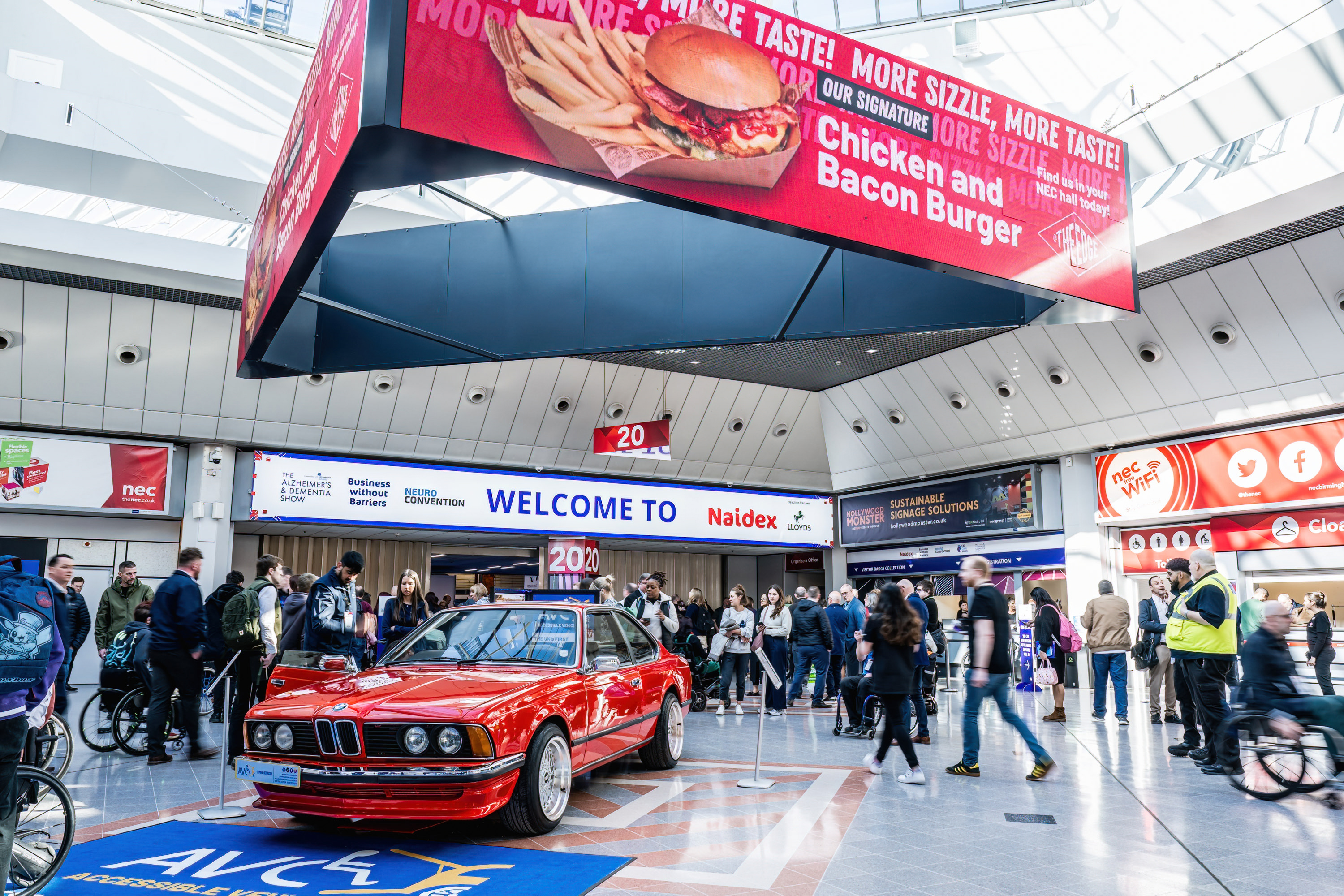 The Naidex entrance with a crowd of people outside. There is a red car outside the entrance and a large hanging banner advertising food at the NEC. 