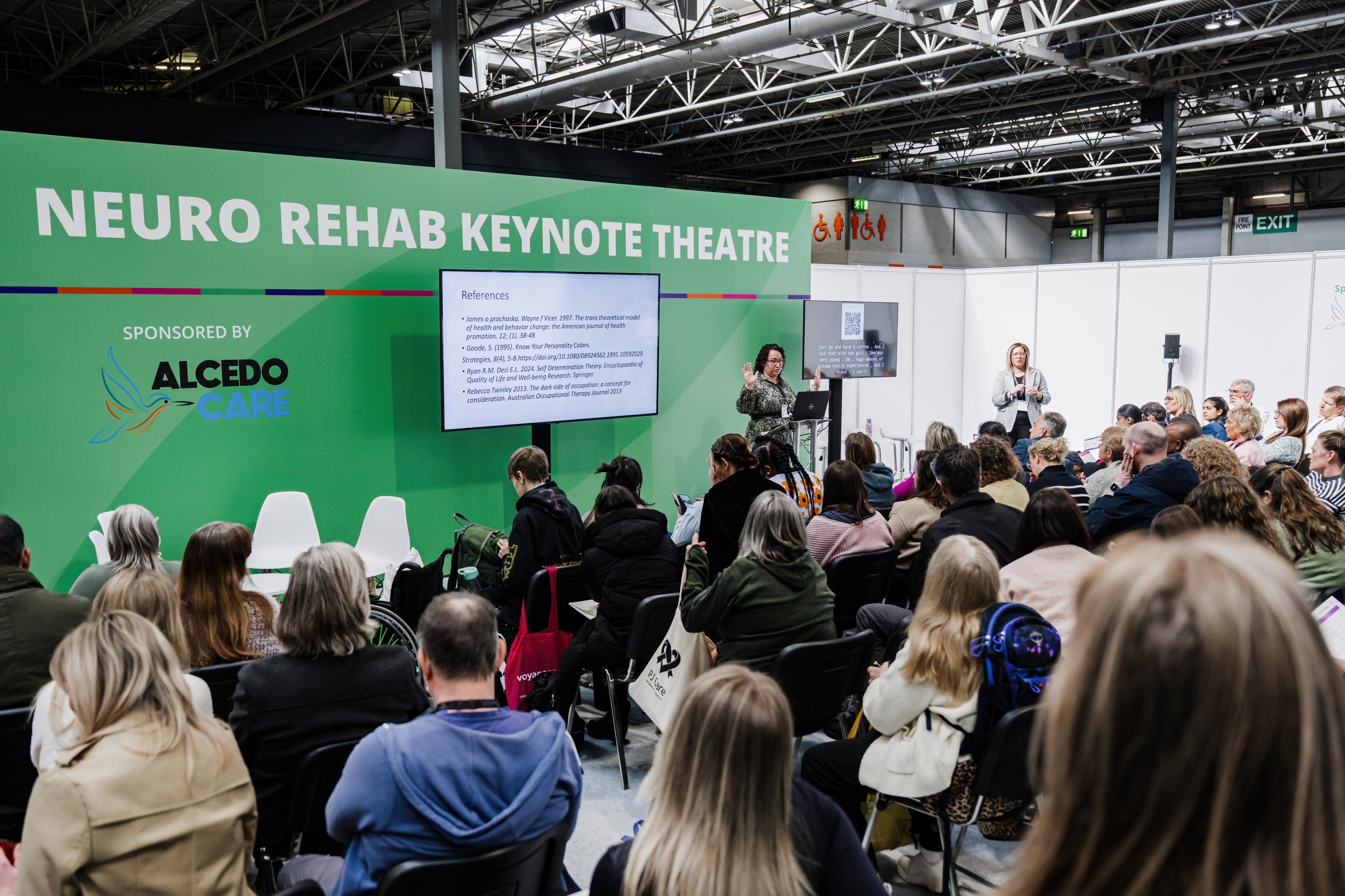 A speaker presents at the Neuro Rehab Keynote Theatre, sponsored by Alcedo Care, standing beside a screen showing a references slide. A large seated audience watches from rows of chairs, with a sign language interpreter and a live captioning screen visible to the right of the stage.