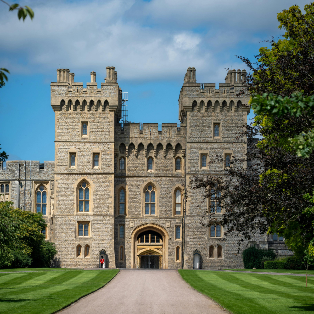 A large historic stone castle with two square towers stands at the end of a long pathway, surrounded by neatly trimmed lawns and trees. A guard in a red uniform stands near the entrance under an arched gateway, with a blue sky and scattered clouds above.