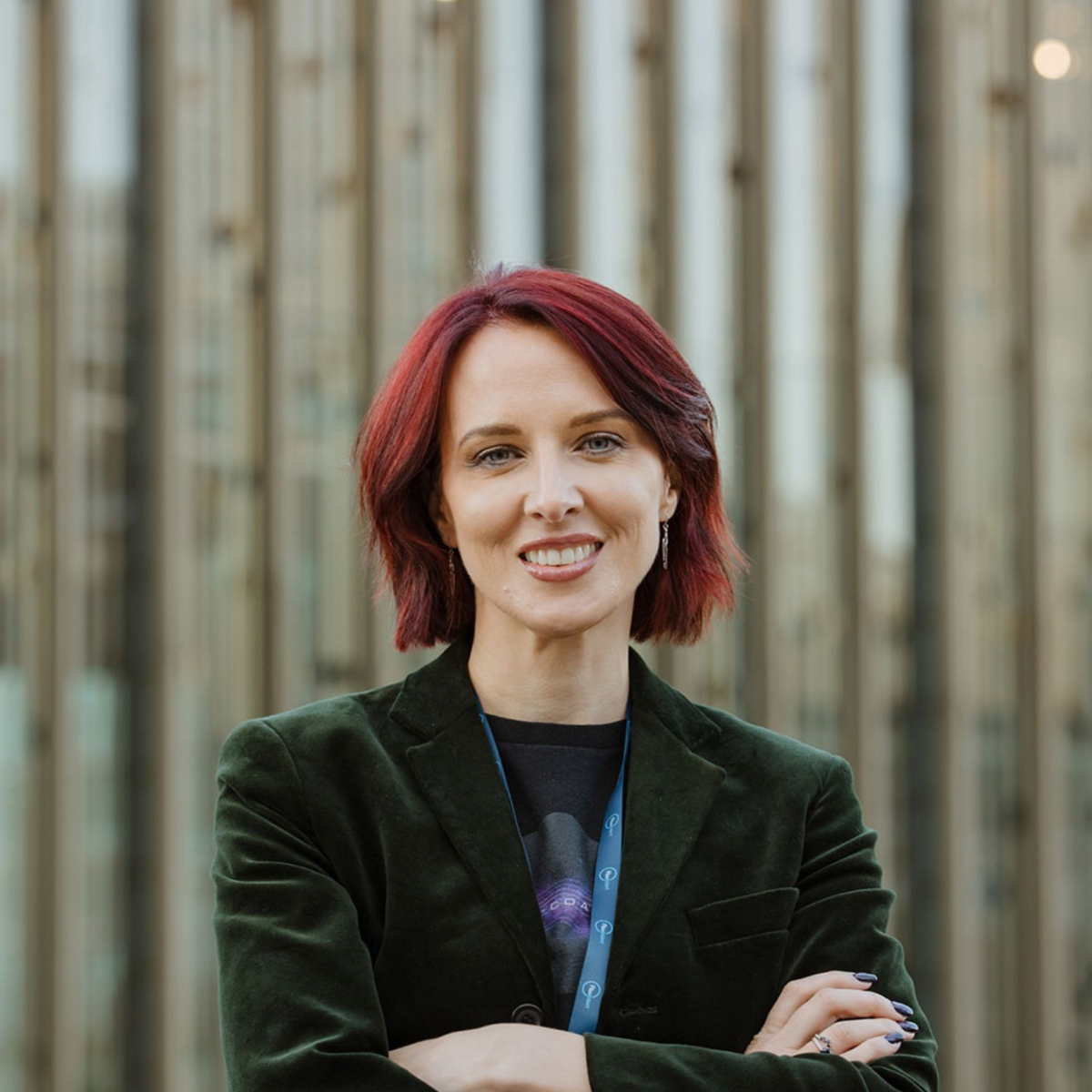 A woman with short red hair smiles at the camera while standing outdoors with her arms crossed. She is wearing a dark green blazer and a lanyard, with a modern building featuring vertical architectural lines blurred in the background.