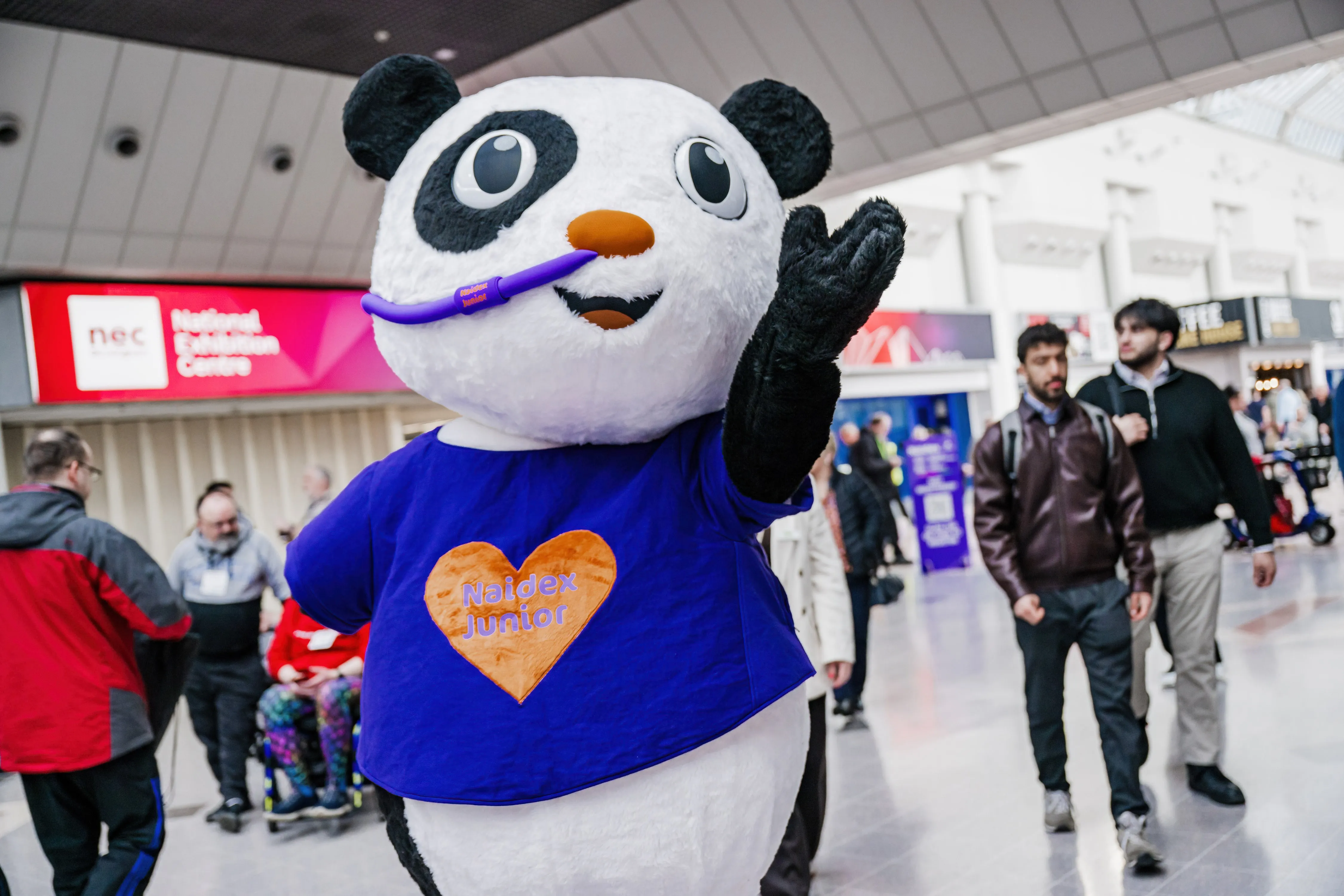 A person in a panda mascot costume wearing a “Naidex Junior” shirt greets attendees in a busy exhibition hall at the NEC.