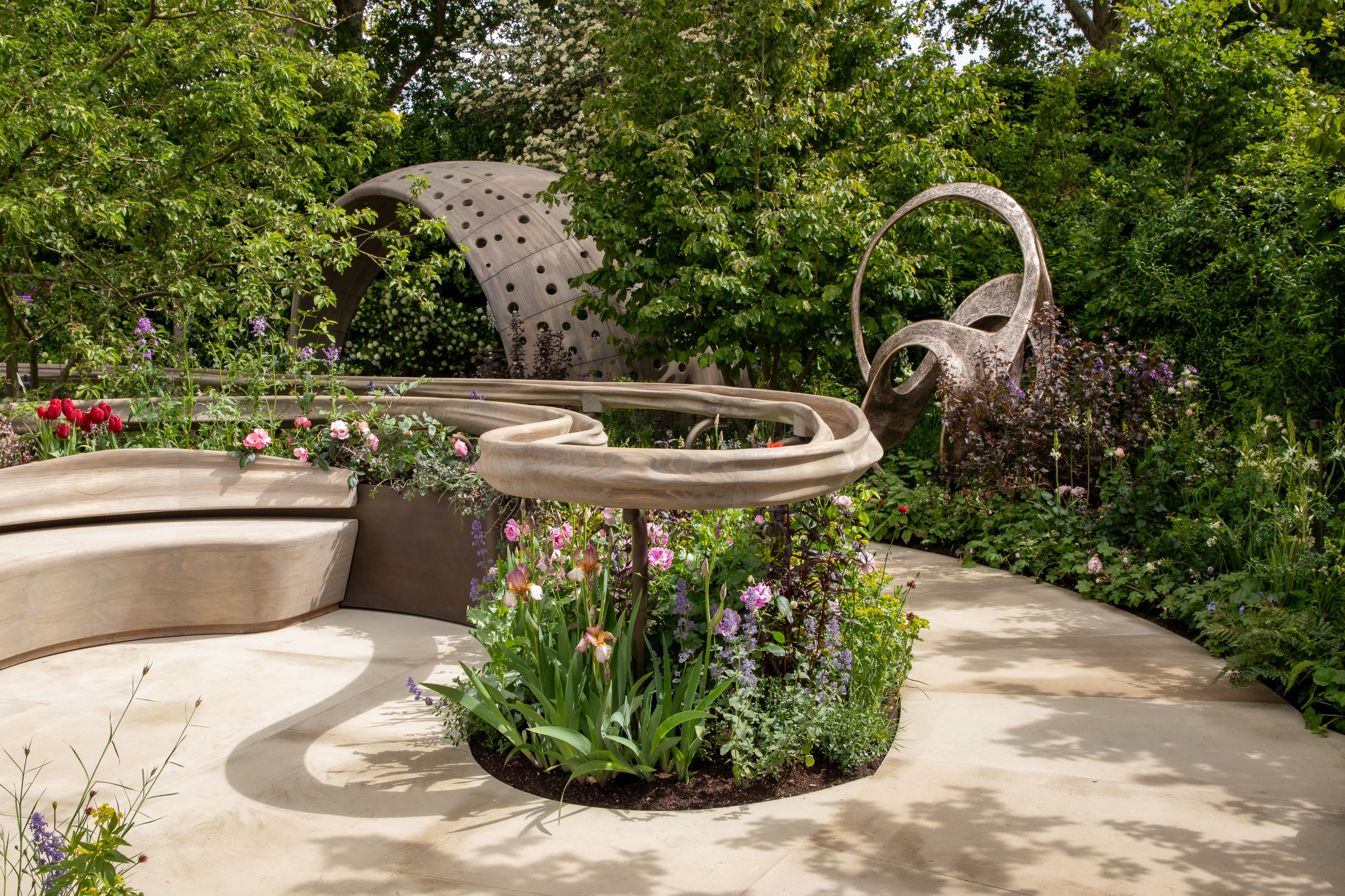 An image of a garden, there are trees in the background and a concrete sculpture in the middle with flowers coming out of it 