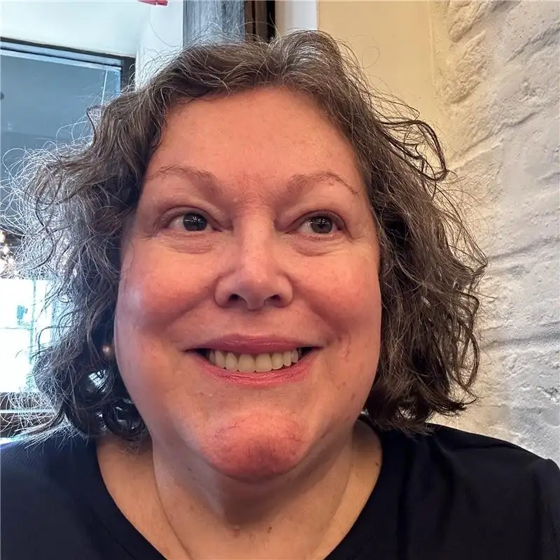A close-up headshot of Ronnie Dean smiling with short, curly hair, wearing a black top. She is looking slightly to the side, with a softly lit indoor background featuring a window and light-coloured wall.