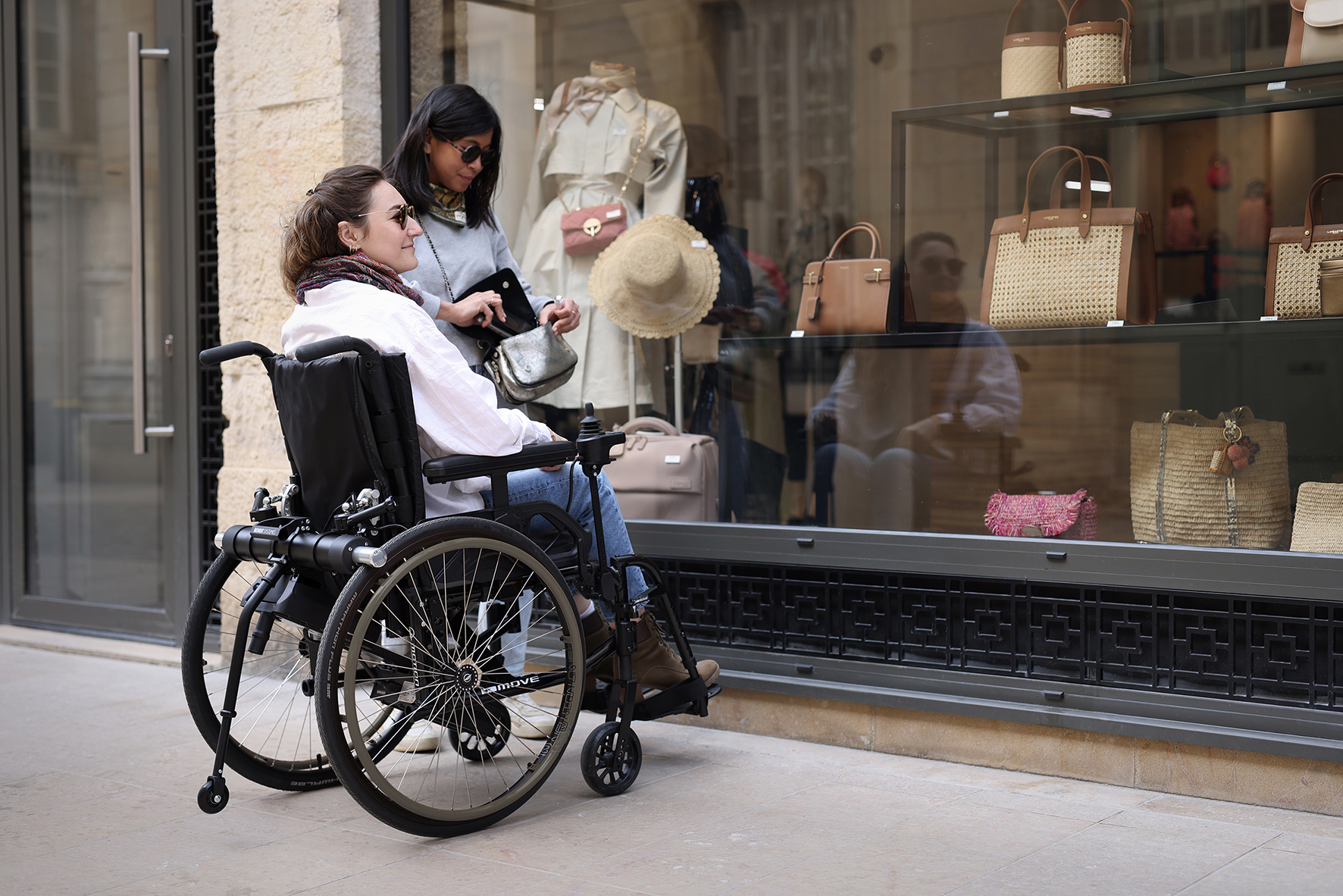 A picture of two women, one is a wheelchair user, looking at a shop window full of handbags