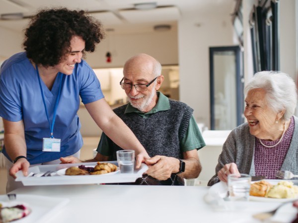 An image of two elderly people sat down smiling as a care worker is bringing over a tray of food 