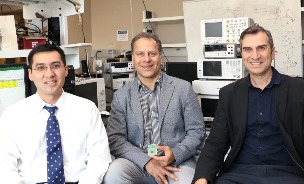 Three men are sitting in a lab.  The man on the left is wearing a white shirt with a blue patterned tie. He has short black hair and glasses.  The man in the middle is wearing a grey blazer over a striped shirt. He has short, curly dark brown hair with some grey. He is holding a small electronic device.  The man on the right is wearing a black blazer over a dark navy shirt. He has short dark brown hair with some grey.