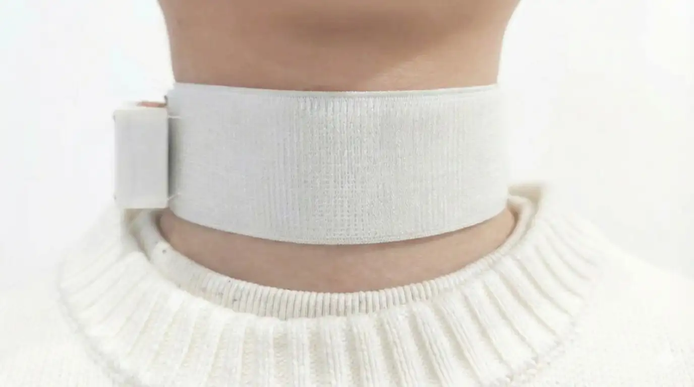 A close-up of a person’s neck wearing a wide white fabric band secured with a fastening on the side, resembling a wearable medical device, Revoice